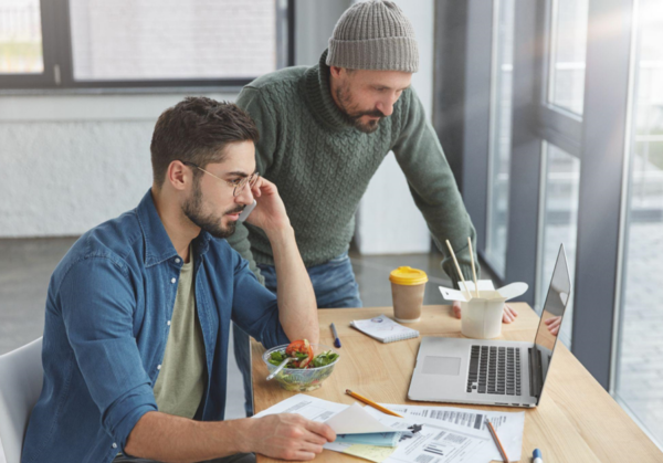 Dwóch mężczyzn pracuje przy drewnianym stole w nowoczesnym biurze. Jeden siedzi przed laptopem, trzyma w ręku dokumenty i rozmawia przez telefon, obok stoi drugi i pochyla się nad ekranem. Na stole leżą kartki z wykresami, długopis, sałatka w misce oraz kubek kawy na wynos.