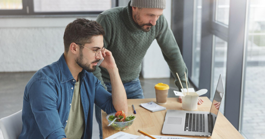 Dwóch mężczyzn pracuje przy drewnianym stole w nowoczesnym biurze. Jeden siedzi przed laptopem, trzyma w ręku dokumenty i rozmawia przez telefon, obok stoi drugi i pochyla się nad ekranem. Na stole leżą kartki z wykresami, długopis, sałatka w misce oraz kubek kawy na wynos.